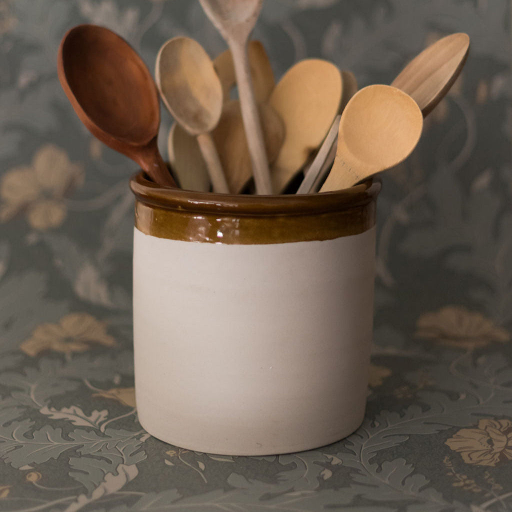 A white Heritage Stoneware crock with a brown rim filled with wooden spoons, set against a patterned background.