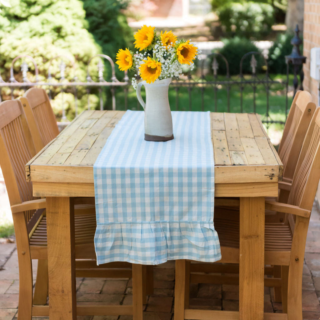 A blue and white gingham table runner with a flirty ruffle is laid out on a wooden dining table outdoors. A vase with yellow flowers adds a complementing touch to the setup.